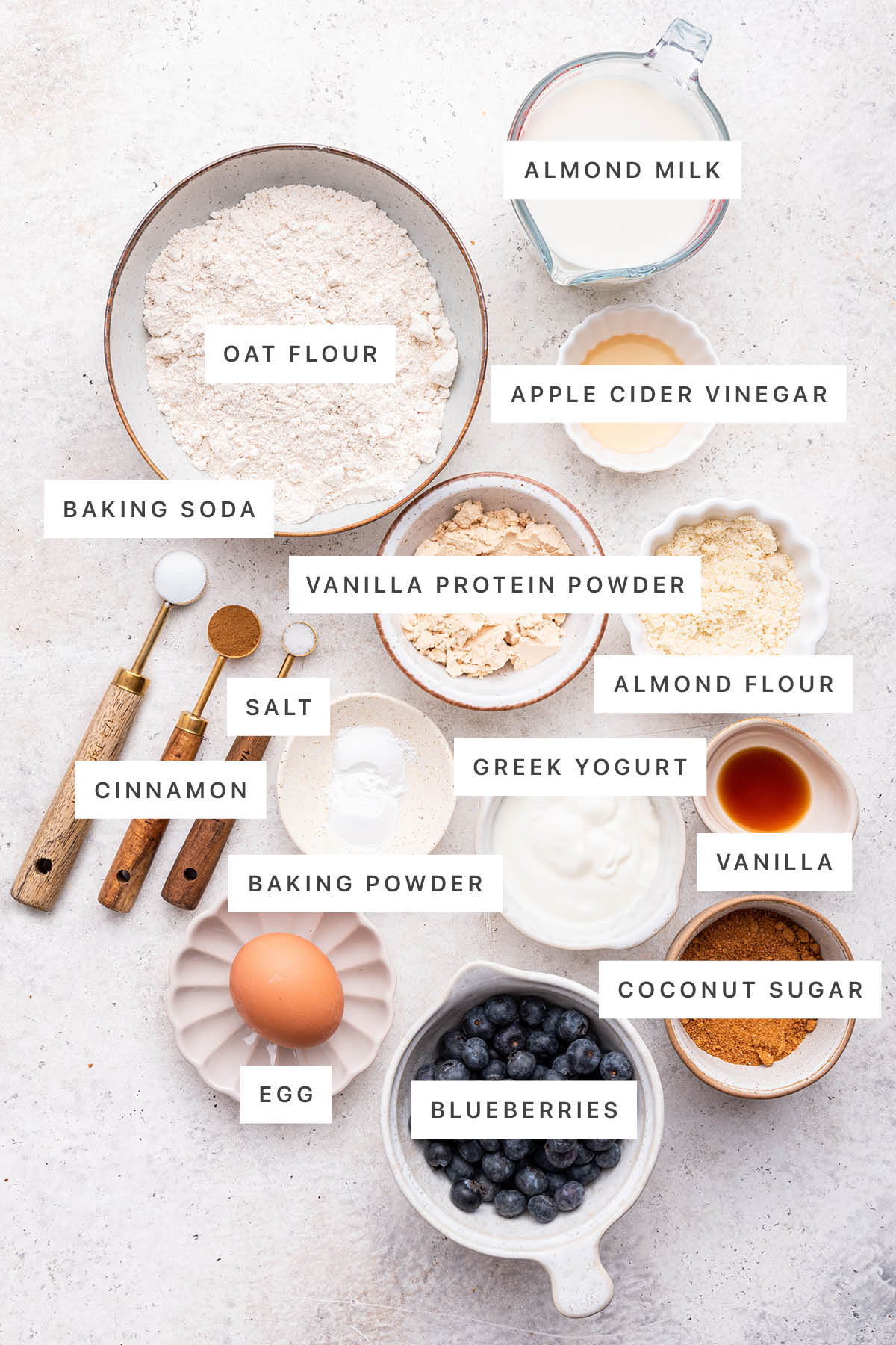 Ingredients measured out to make Blueberry Protein Muffins: oat flour, almond milk, apple cider vinegar, baking soda, vanilla protein powder, salt, almond flour, cinnamon, Greek yogurt, baking powder, vanilla, egg, blueberries and coconut sugar.
