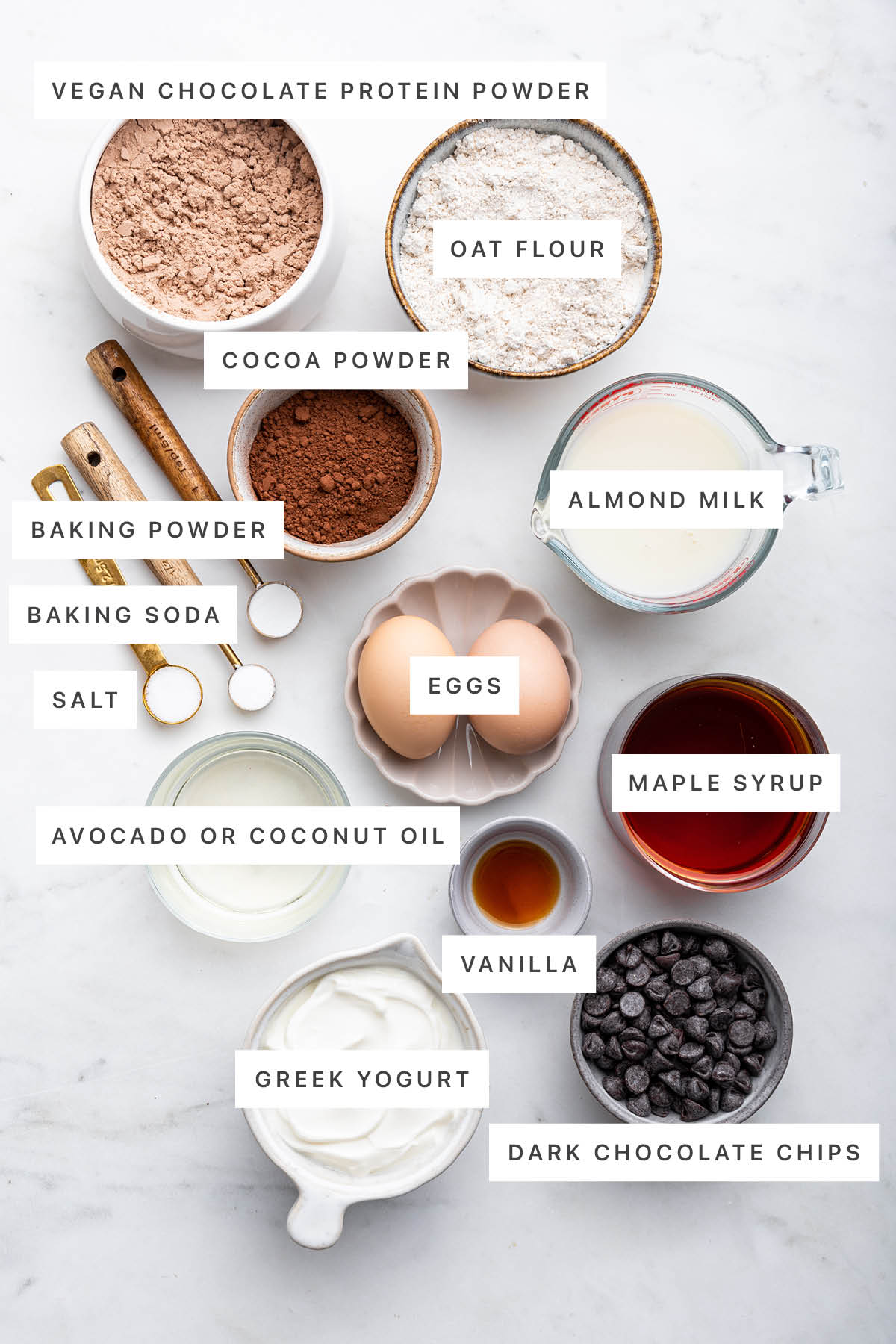 Ingredients measured out to make Protein Cake: vegan chocolate protein powder, oat flour, cocoa powder, almond milk, baking powder, baking soda, salt, eggs, maple syrup, coconut oil, vanilla, Greek yogurt and dark chocolate chips.