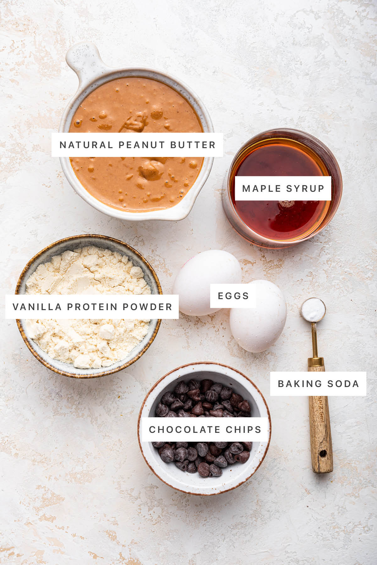 Ingredients measured out to make Protein Cookies: natural peanut butter, maple syrup, vanilla protein powder, eggs, chocolate chips and baking soda.