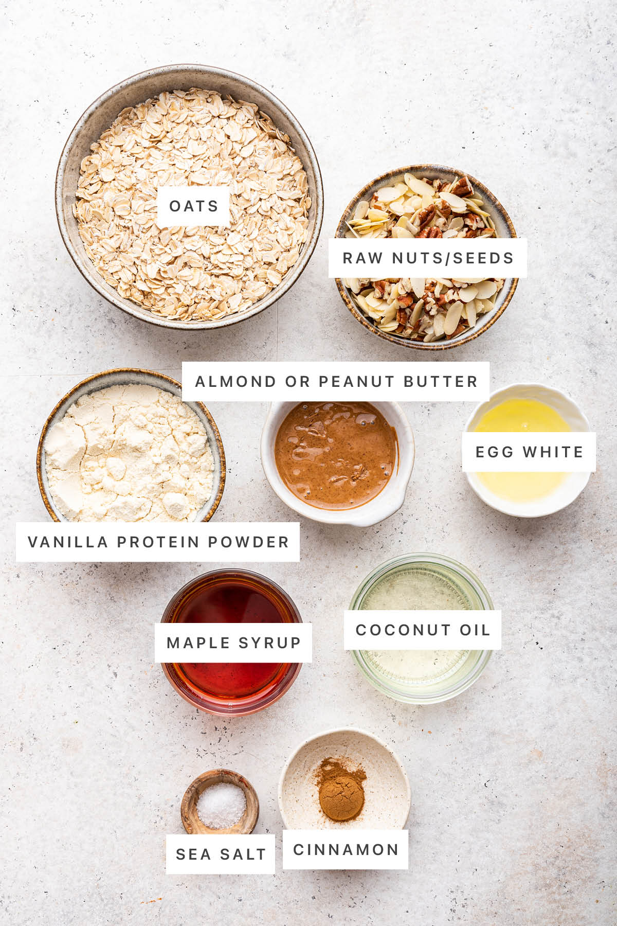 Ingredients measured out to make protein granola: oats, raw nuts/seeds, almond or peanut butter, egg white, vanilla protein powder, maple syrup, coconut oil, sea salt and cinnamon.