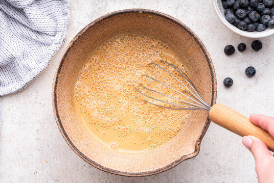 A woman's hand uses a whisk to stir the ingredients in a large mixing bowl used for blueberry protein muffins.