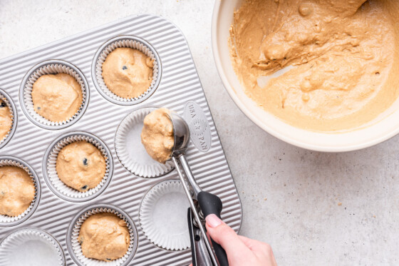 A woman's hand uses a cookie scoop to measure out the blueberry protein muffins in a muffin tin.