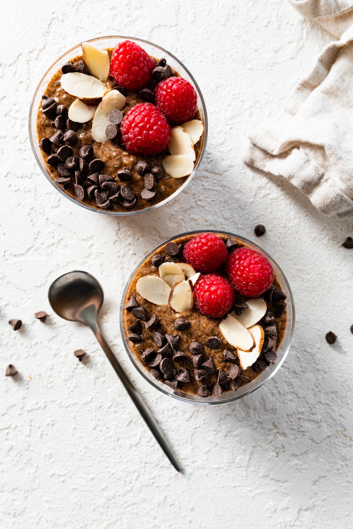 Two chocolate chia puddings in glass cups topped with chocolate chips, sliced almonds, and fresh raspberries.