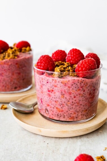 A raspberry chia pudding in a glass cup, topped with fresh raspberries and granola.