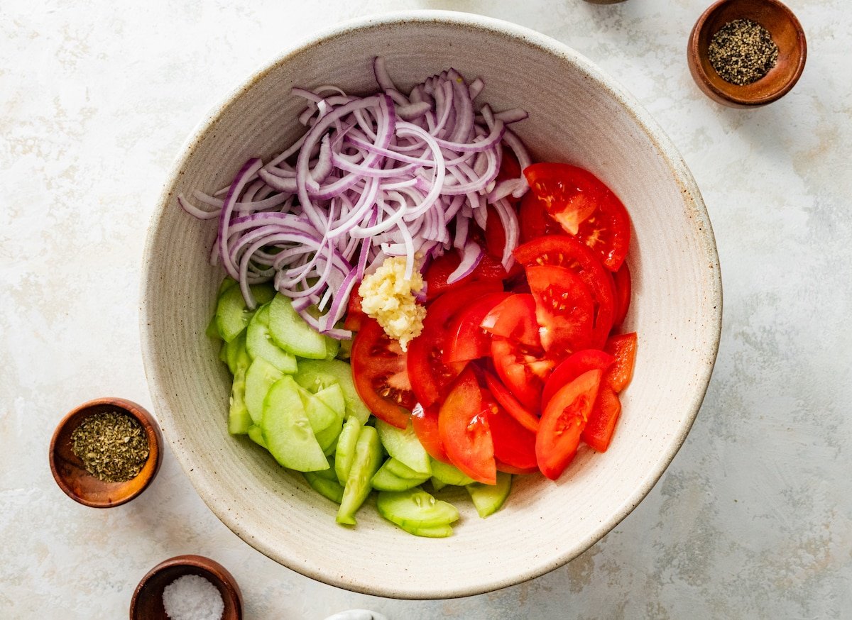 Sliced cucumber, tomato, red onion, and garlic in a large mixing bowl.