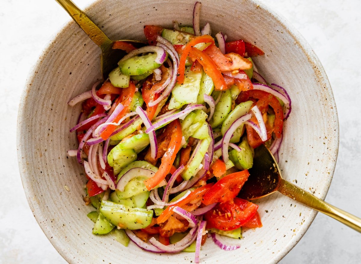 Sliced cucumber, tomato, red onion, and garlic in a large mixing bowl with two serving spoons.