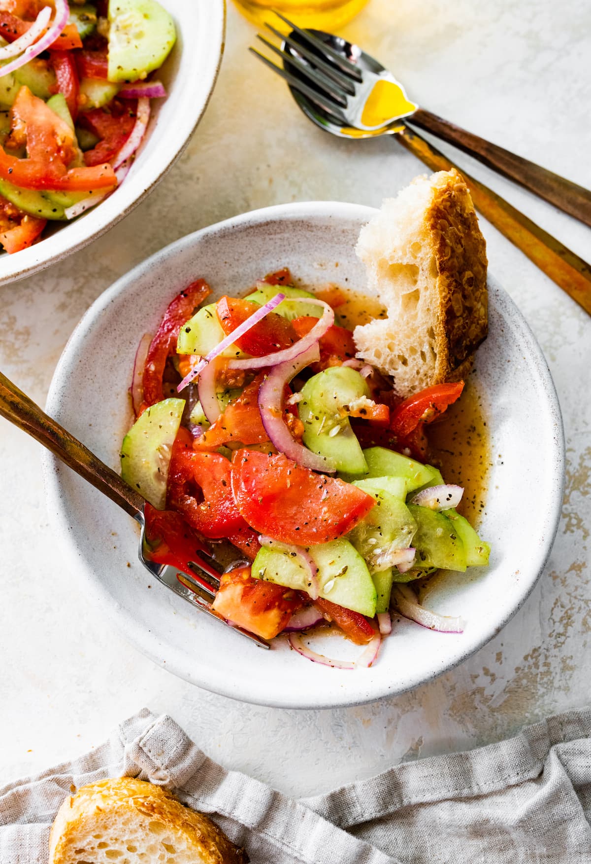In a small white bowl, a cucumber tomato salad with sliced red onion, salt, pepper, and a slice of bread.