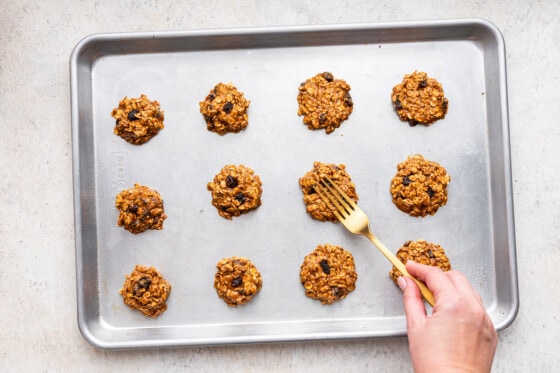 A woman's hand uses a fork to press down oatmeal raisin cookies on a baking tray.