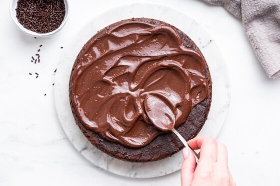 A chocolate frosting is spread on a protein cake with a metal spoon.