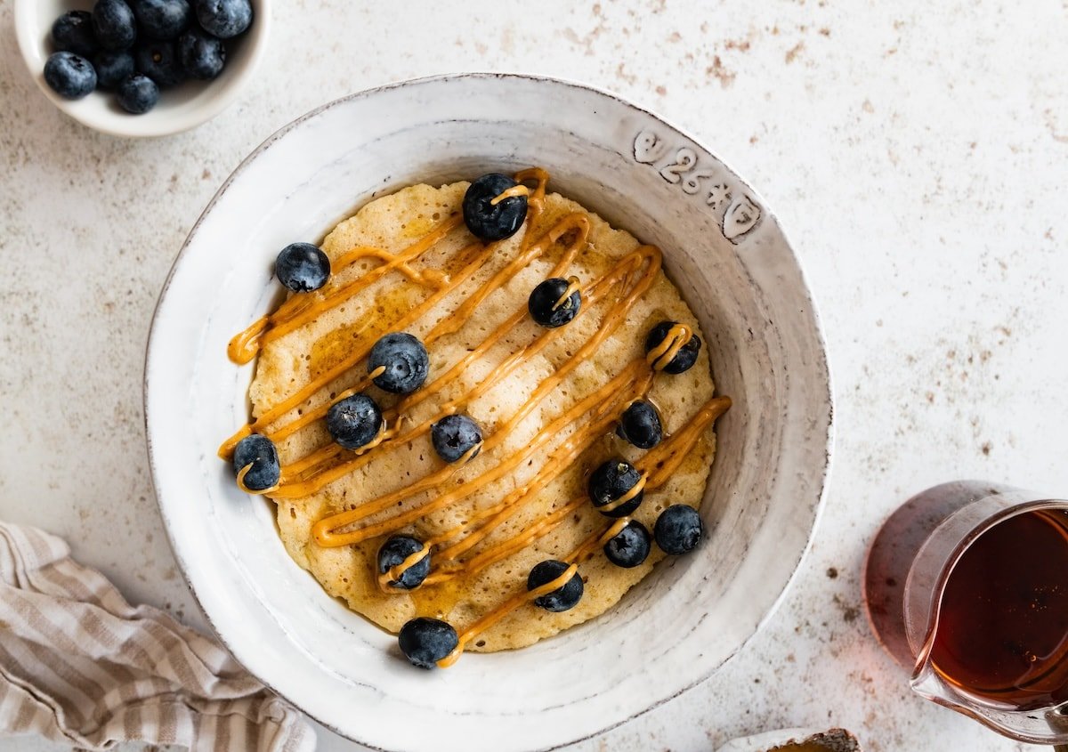 A protein pancake cooked in a bowl topped with fresh blueberries and a drizzle of peanut butter.