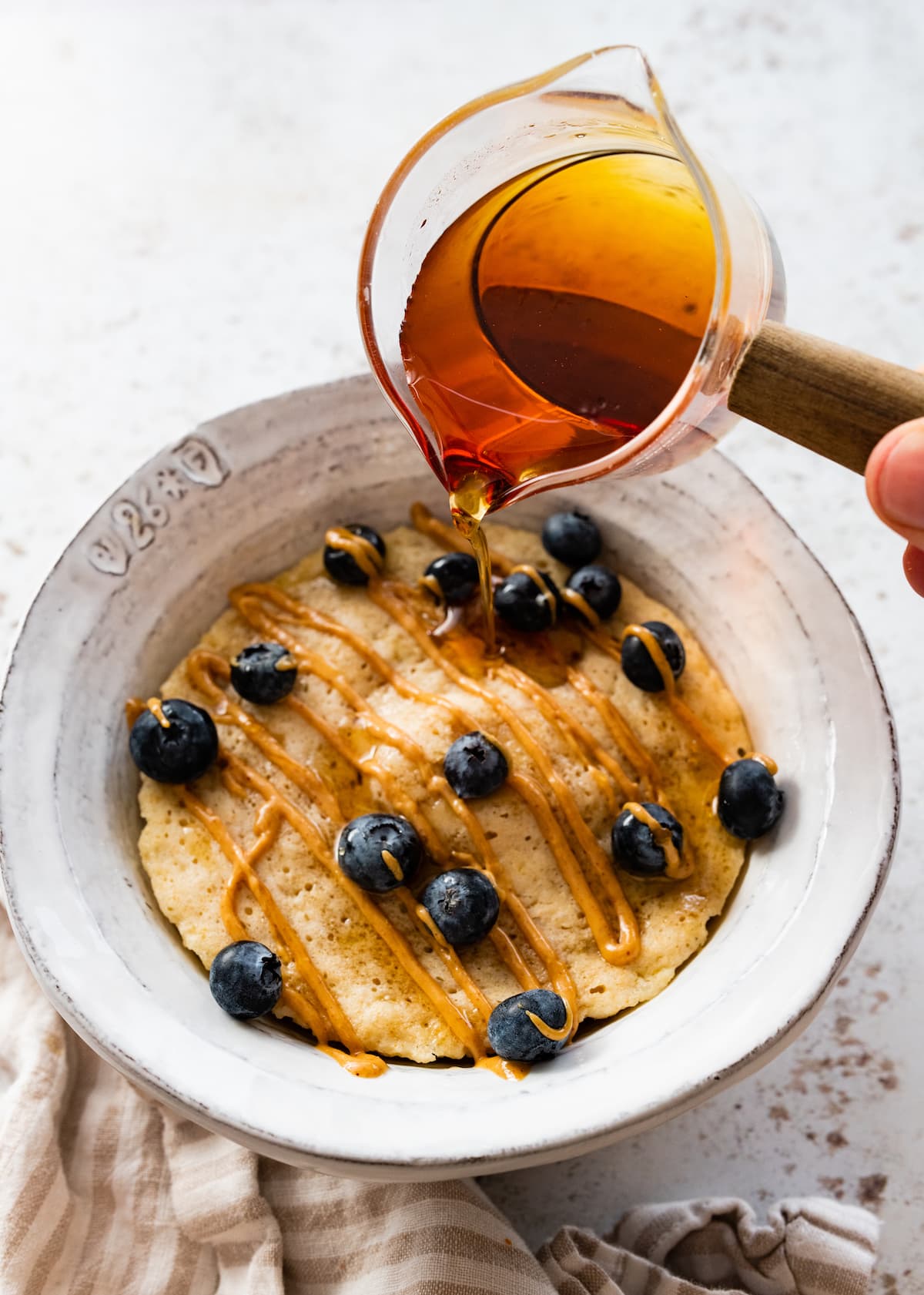 A protein pancake in a bowl topped with a drizzle of peanut butter and fresh blueberries being topped off with some maple syrup.
