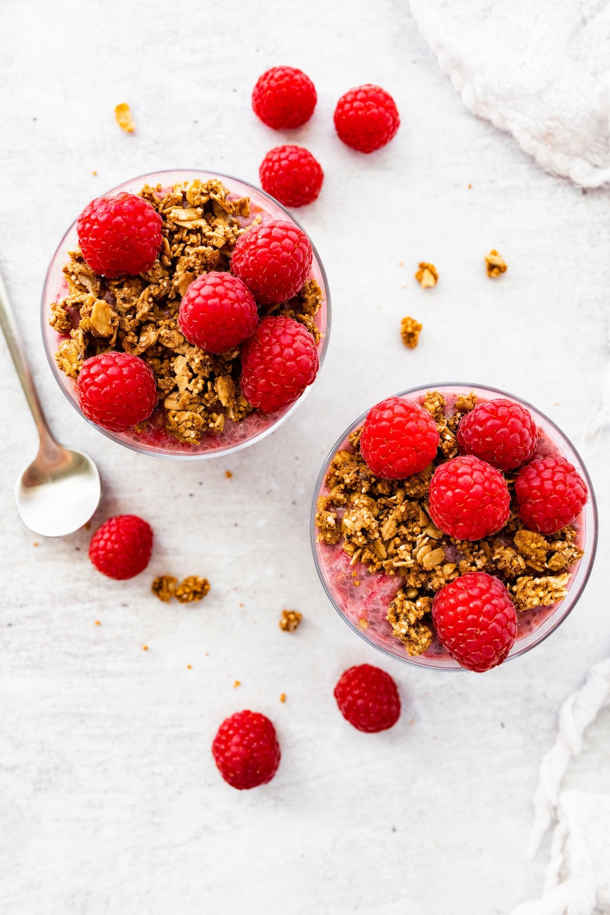 Two raspberry chia puddings in glass cups, topped with fresh raspberries and granola.