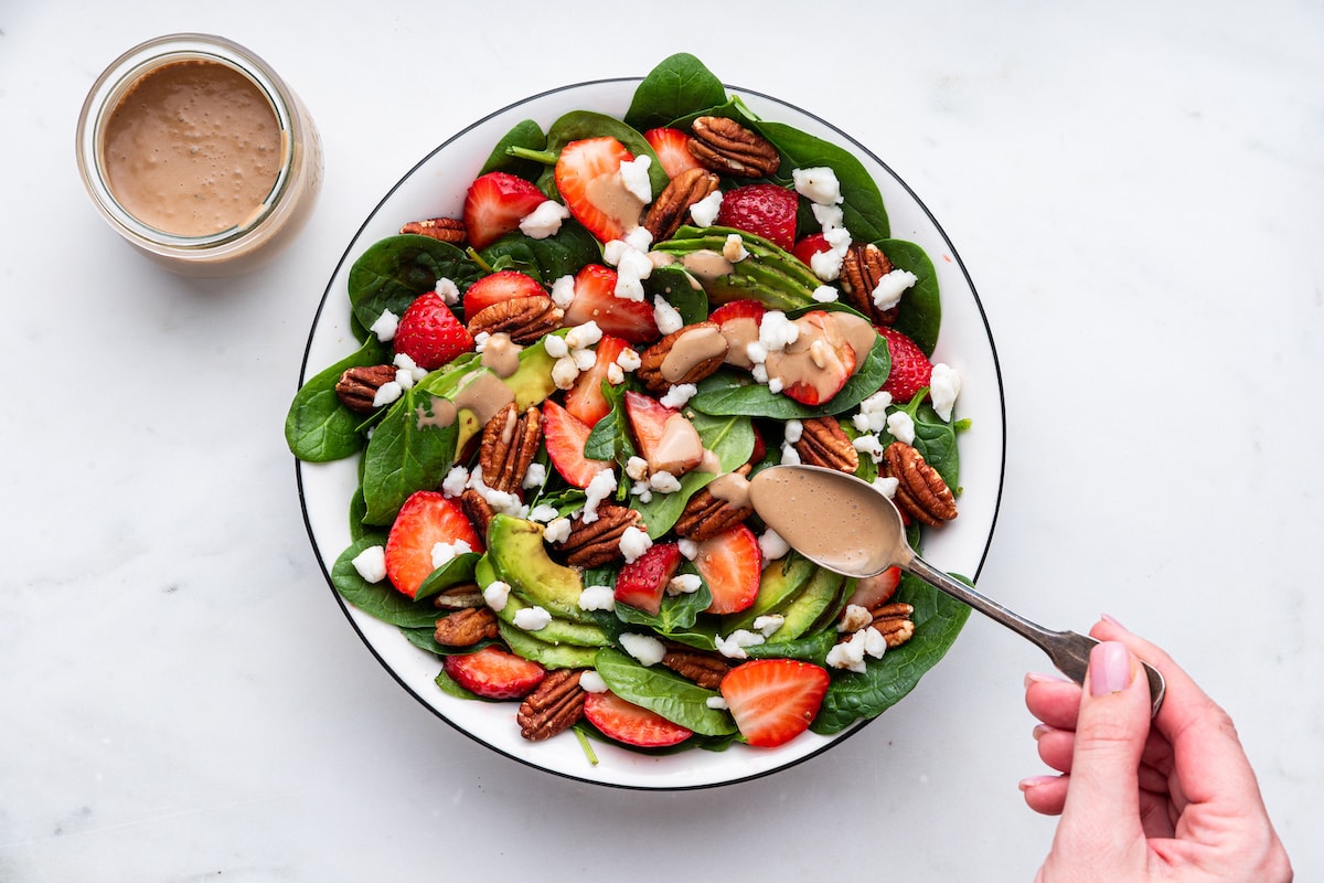 A woman's hand drizzling balsamic dressing on top of a strawberry spinach salad.