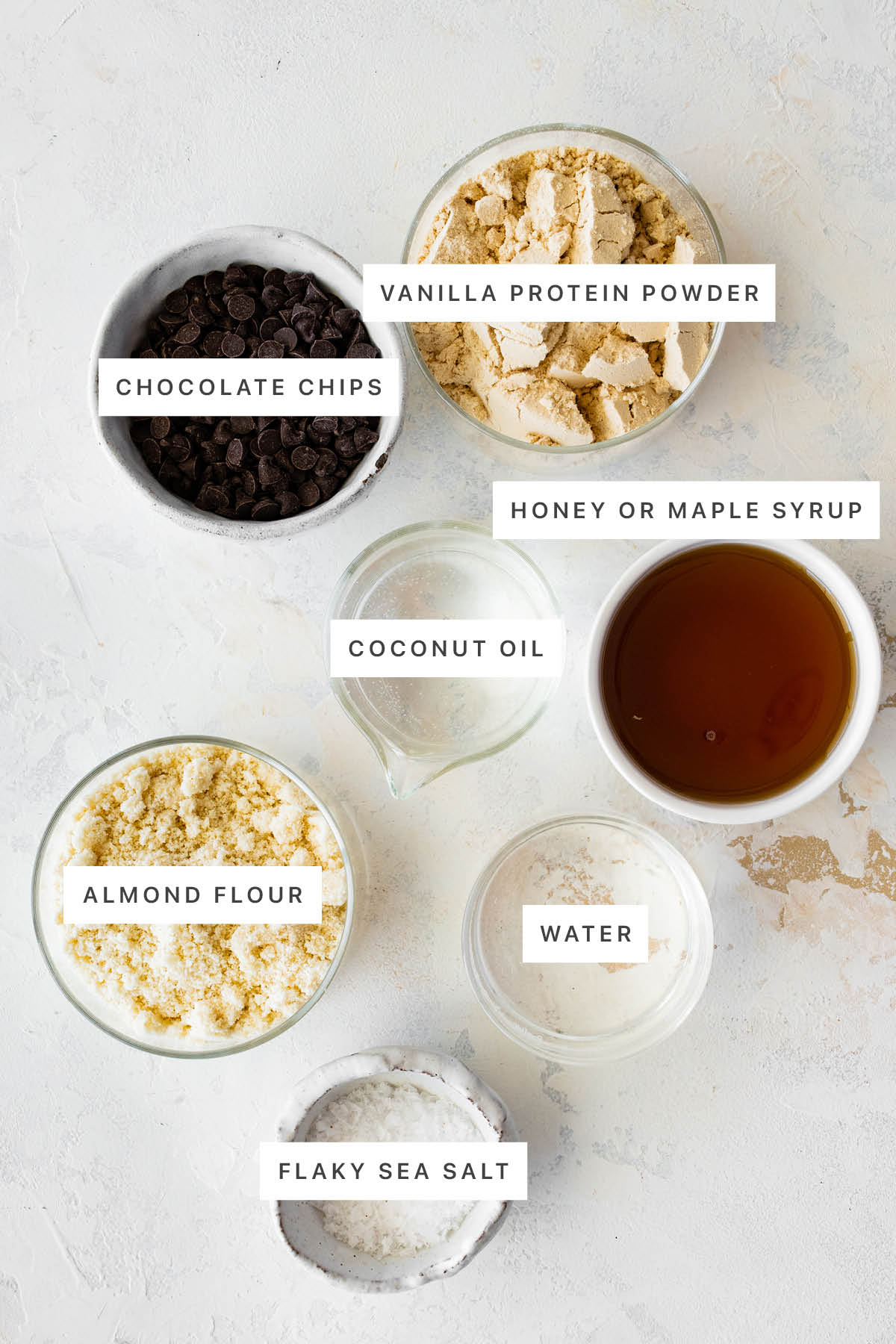 Ingredients measured out to make Protein Cookie Dough Bark: chocolate chips, vanilla protein powder, maple syrup or honey, coconut oil, almond flour, water and flaky sea salt.