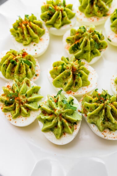 Avocado deviled eggs near one another on a plate and topped with a dash of paprika and fresh herbs.