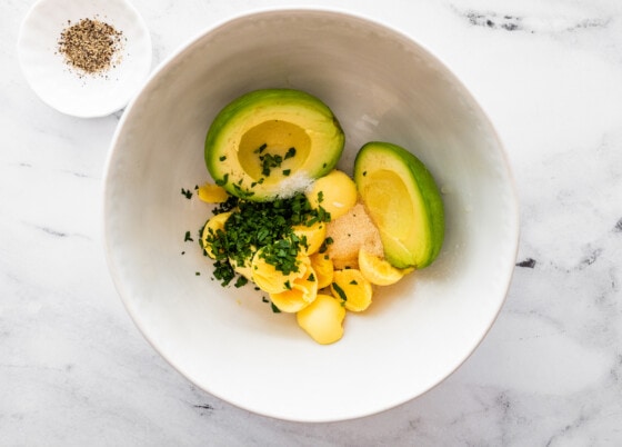 Hard boiled egg yolks, an avocado, fresh herbs, and seasonings in a large mixing bowl.