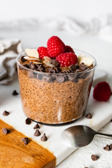 A chocolate chia pudding in a glass cup topped with chocolate chips, sliced almonds, and fresh raspberries.
