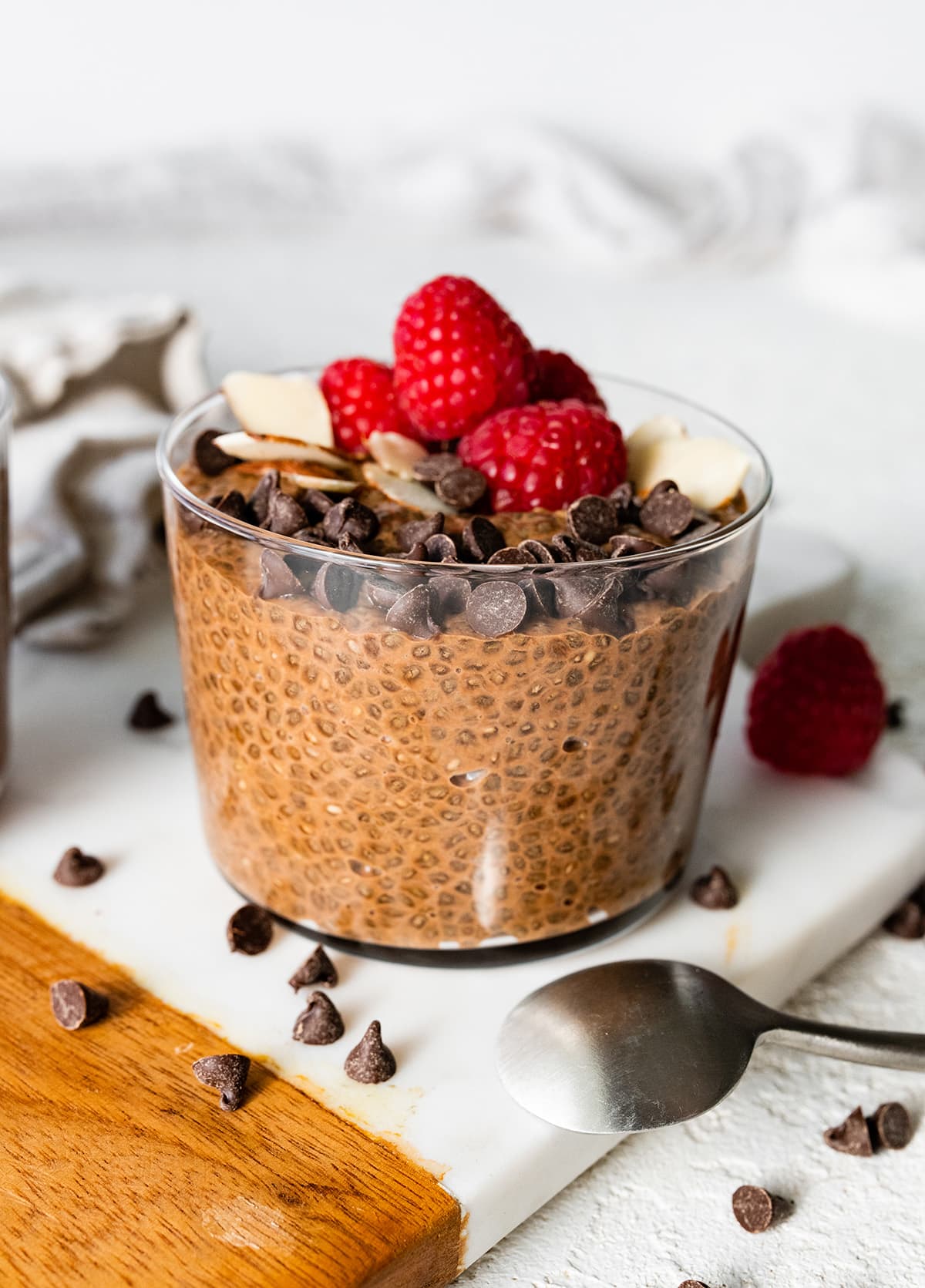 A chocolate chia pudding in a glass cup topped with chocolate chips, sliced almonds, and fresh raspberries.