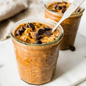 2 chocolate overnight oats in mason jars topped with chocolate chips and a drizzle of peanut butter.