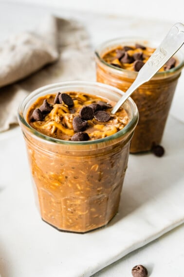 2 chocolate overnight oats in mason jars topped with chocolate chips and a drizzle of peanut butter.