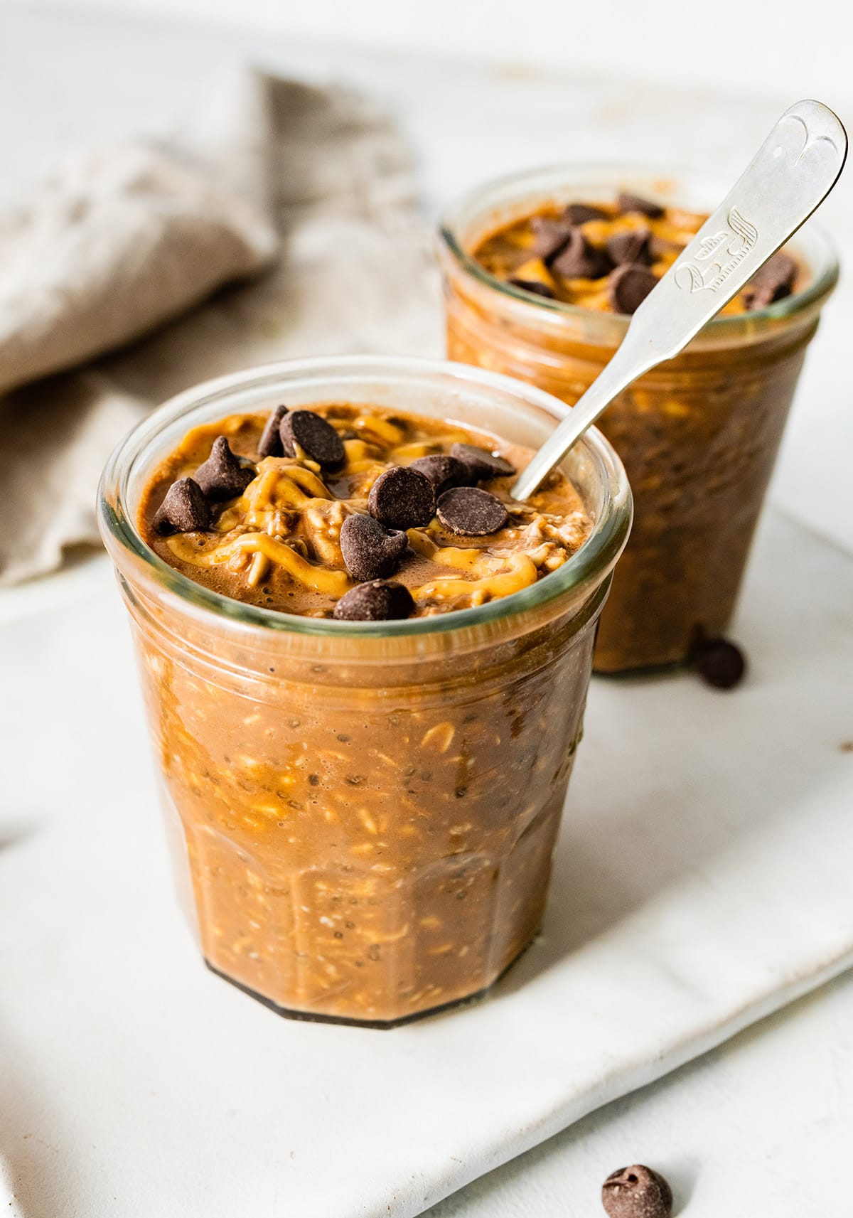 2 chocolate overnight oats in mason jars topped with chocolate chips and a drizzle of peanut butter.