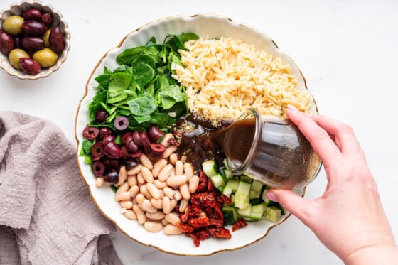 A dressing being poured in a large bowl of ingredients used for a mediterranean orzo salad.