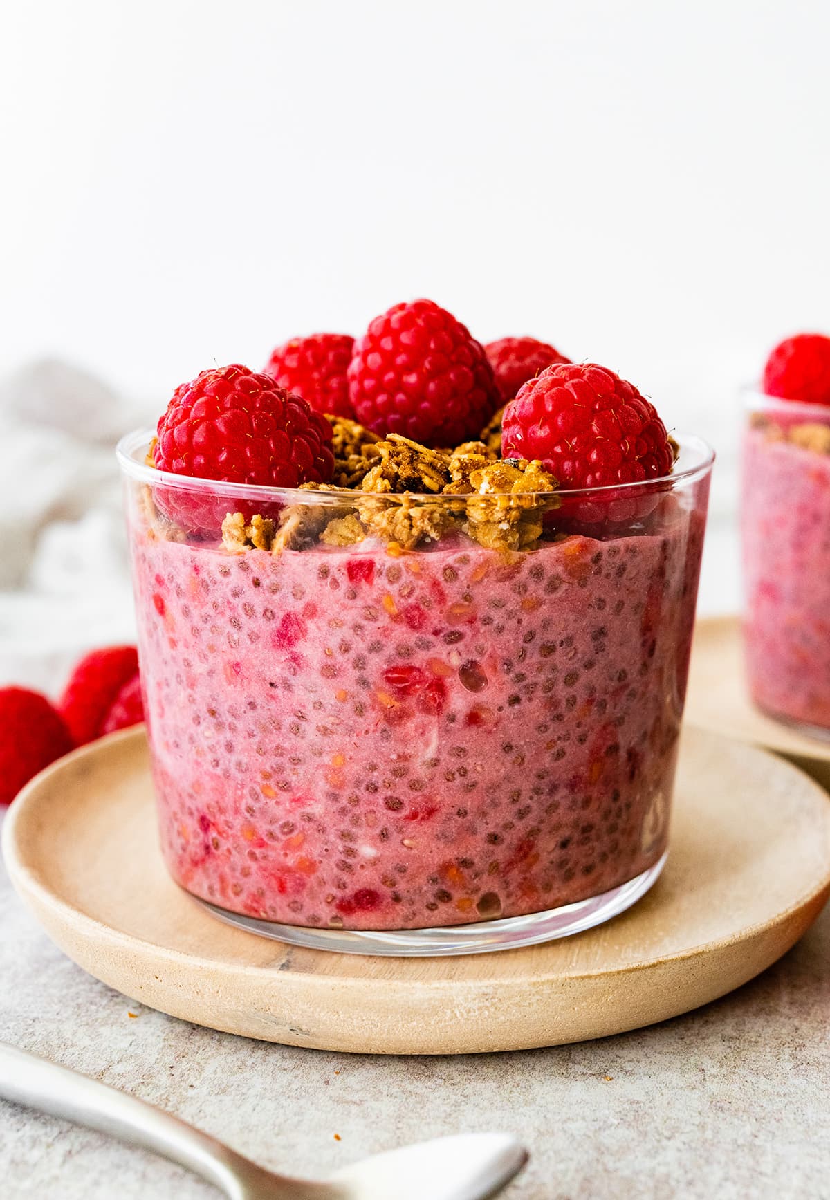 A raspberry chia pudding in a glass cup, topped with fresh raspberries and granola.