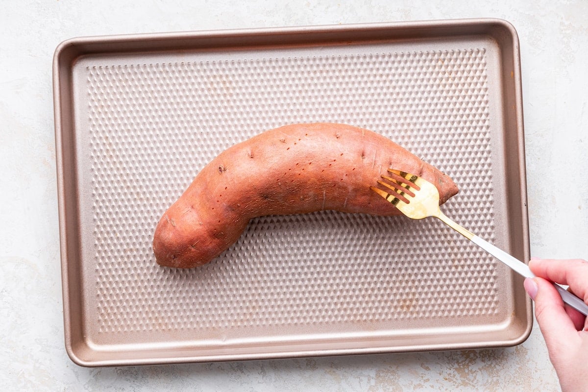 A woman's hand uses a fork to poke holes in a sweet potato on a baking tray.