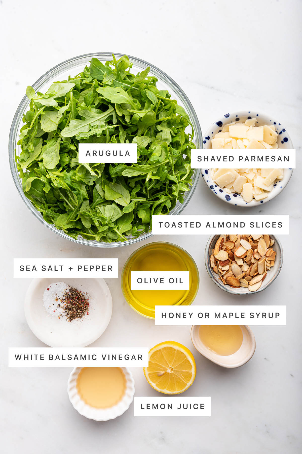 Ingredients measured out to make Arugula Salad: arugula, shaved parmesan, toasted almond slices, sea salt, pepper, olive oil, honey, white balsamic vinegar and lemon juice.