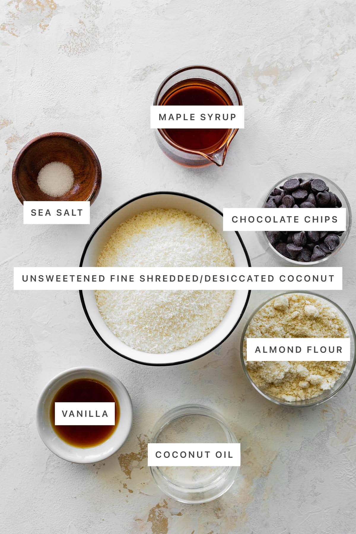 Ingredients measured out to make Coconut Macaroons: sea salt, maple syrup, chocolate chips, unsweetened desiccated coconut, almond flour, vanilla and coconut oil.