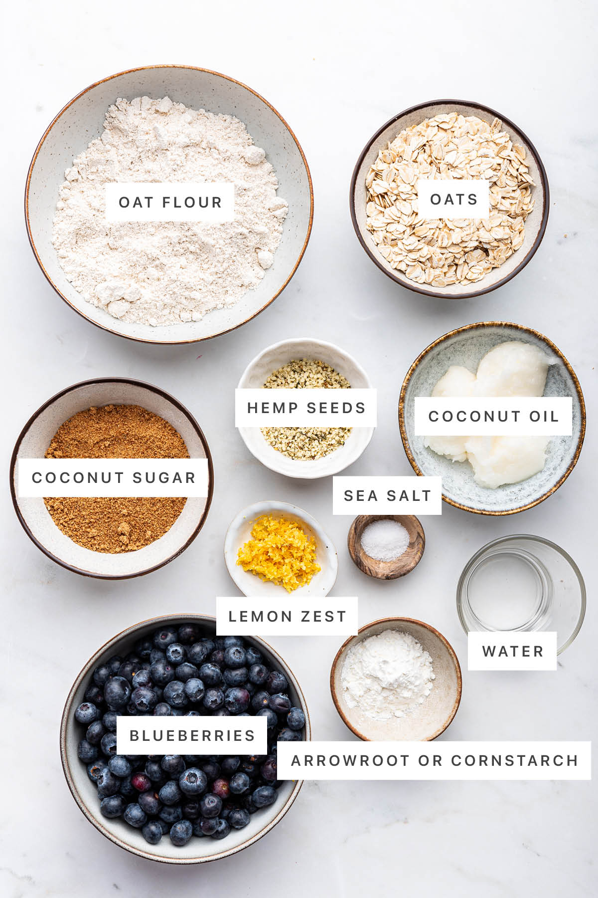 Ingredients measured out to make Healthy Blueberry Crumble Bars: oat flour, oats, hemp seeds, coconut oil, coconut sugar, sea salt, lemon zest, water, blueberries and arrowroot powder.