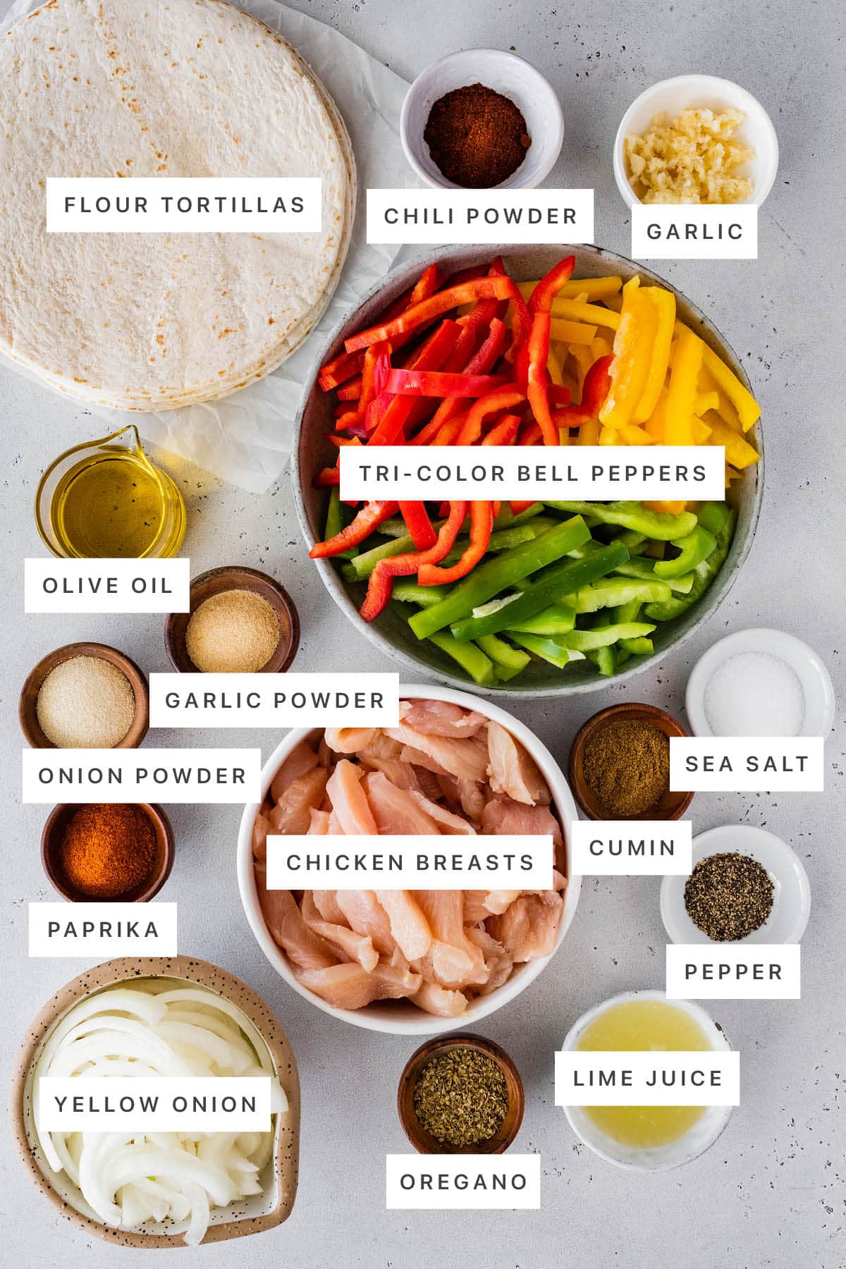 Ingredients measured out to make Healthy Chicken Fajitas: flour tortillas, chili powder, garlic, tri-colored peppers, olive oil, garlic powder, onion powder, paprika, chicken breasts, cumin, sea salt, pepper, lime juice, oregano and yellow onion.