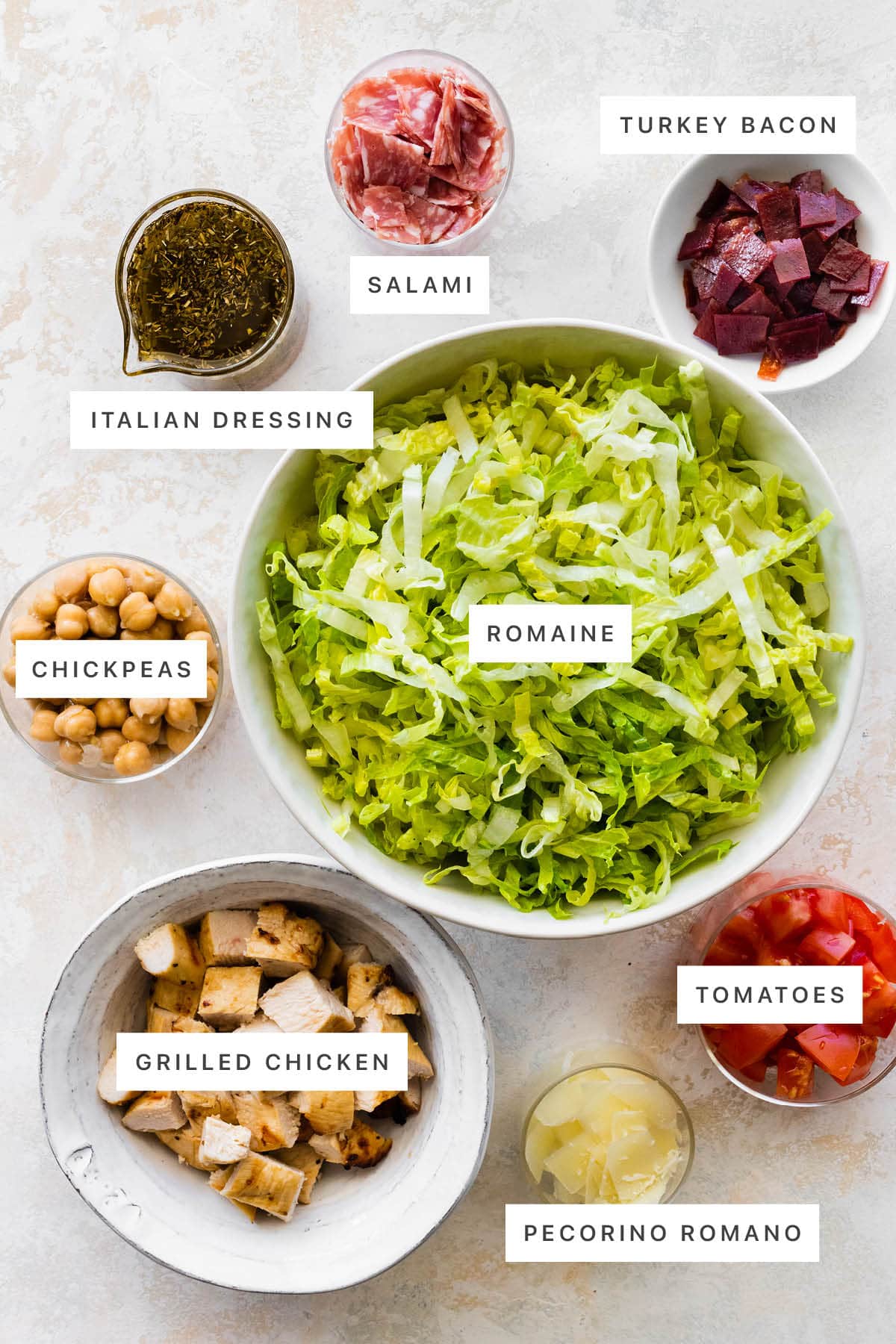 Ingredients measured out to make Jennifer Aniston Cobb Salad: Italian dressing, salami, turkey bacon, romaine, chickpeas, grilled chicken, pecorino romano and tomatoes.