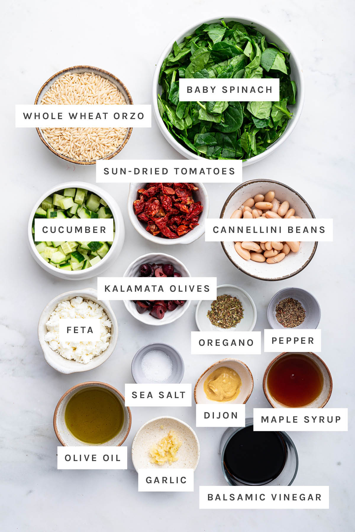 Ingredients measured out to make Mediterranean Orzo Salad: whole wheat orzo, baby spinach, sun-dried tomatoes, cucumber, cannellini beans, kalamata olives, feta, sea salt, oregano, pepper, olive oil, garlic, maple syrup and balsamic vinegar.