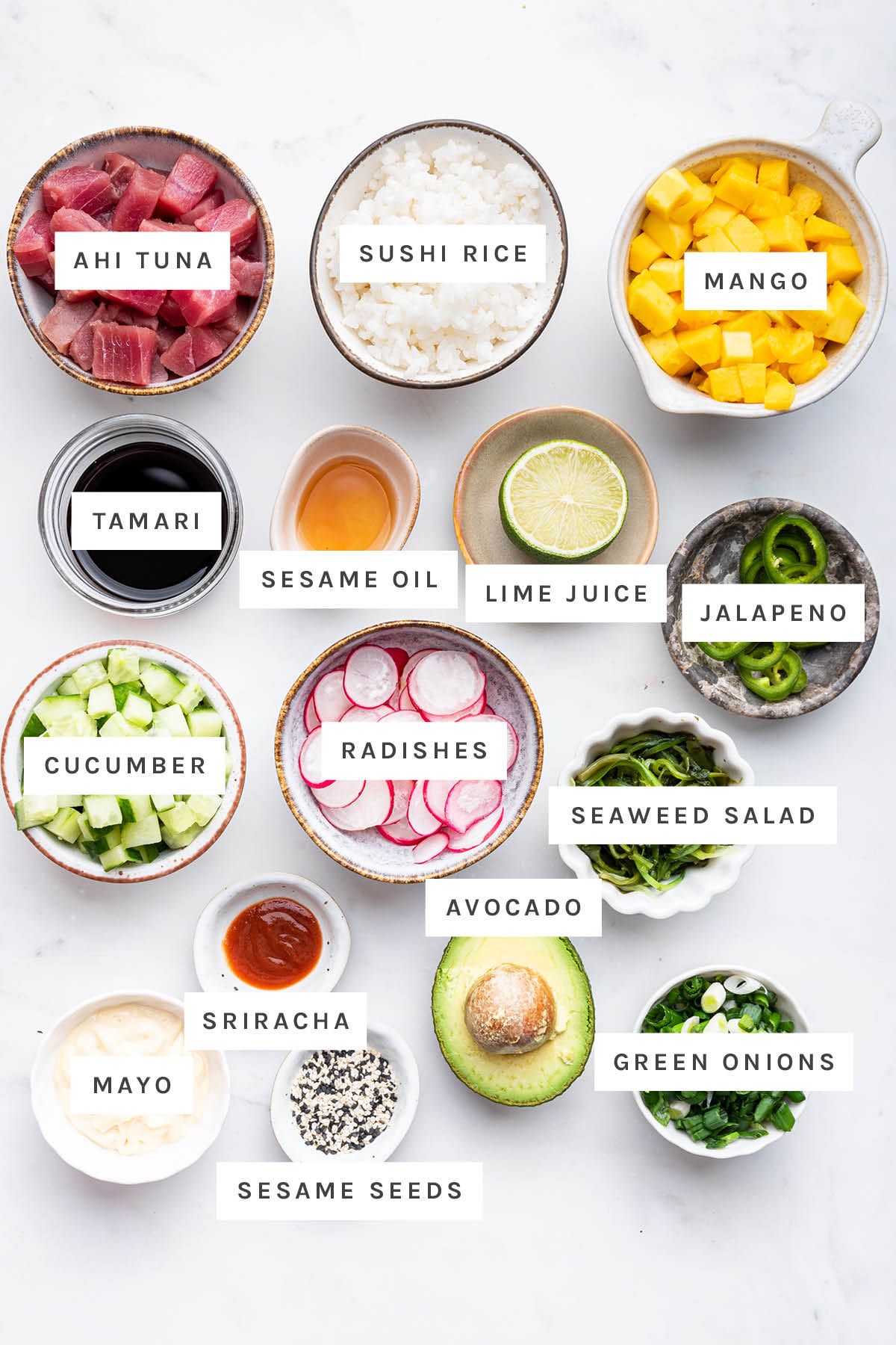 Ingredients measured out to make poke bowls: ahi tuna, sushi rice, mango, jalapeno, seaweed salad, green onions, radishes, sesame oil cucumber, avocado, black sesame seeds, lime, tamari and sriracha mayo.