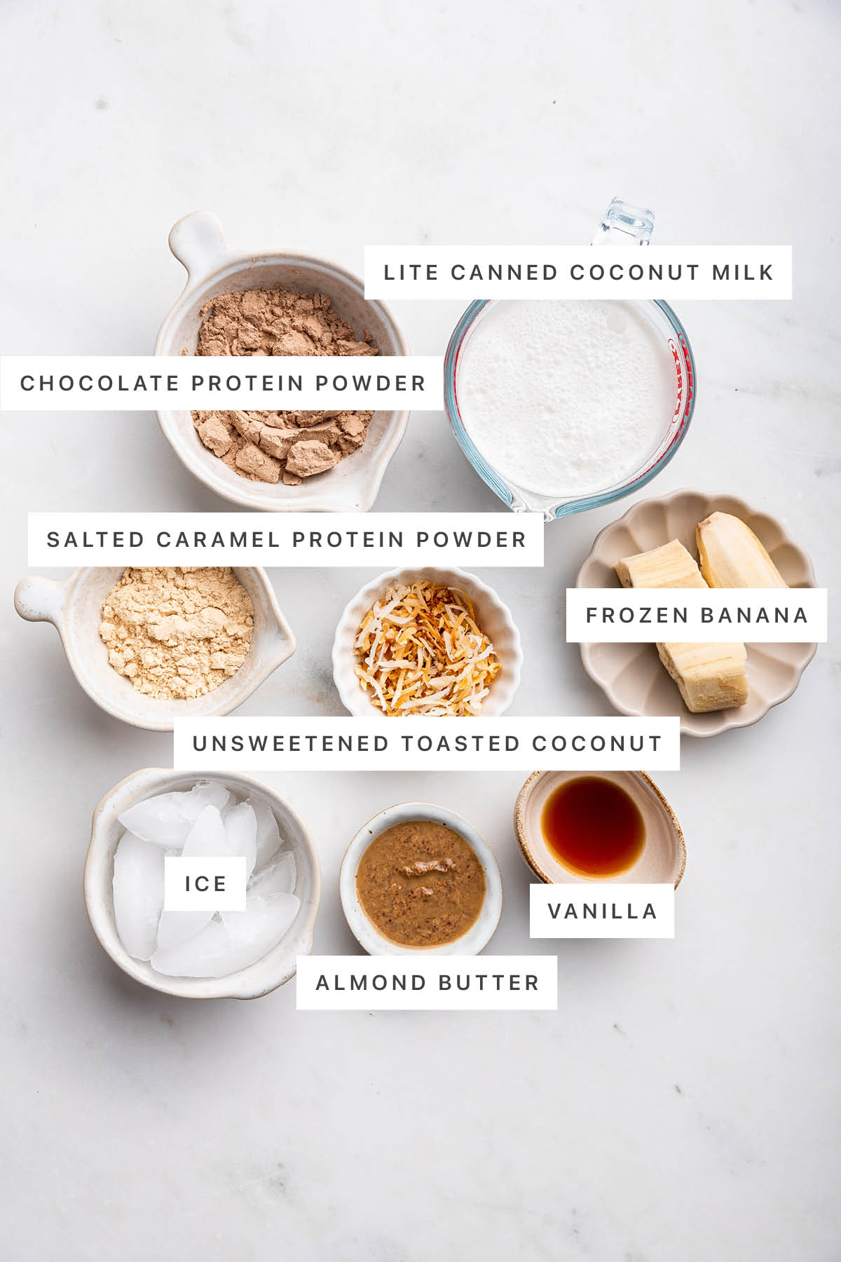Ingredients measured out to make Samoa Protein Shake: chocolate protein powder, lite canned coconut milk, salted caramel protein powder, frozen banana, unsweetened toasted coconut, ice, almond butter and vanilla.
