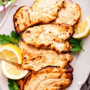 Five cooked chicken cutlets on a serving plate served with lemon wedges and fresh parsley.