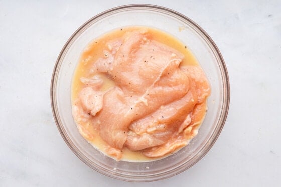 Raw chicken cutlets marinating in a glass mixing bowl.