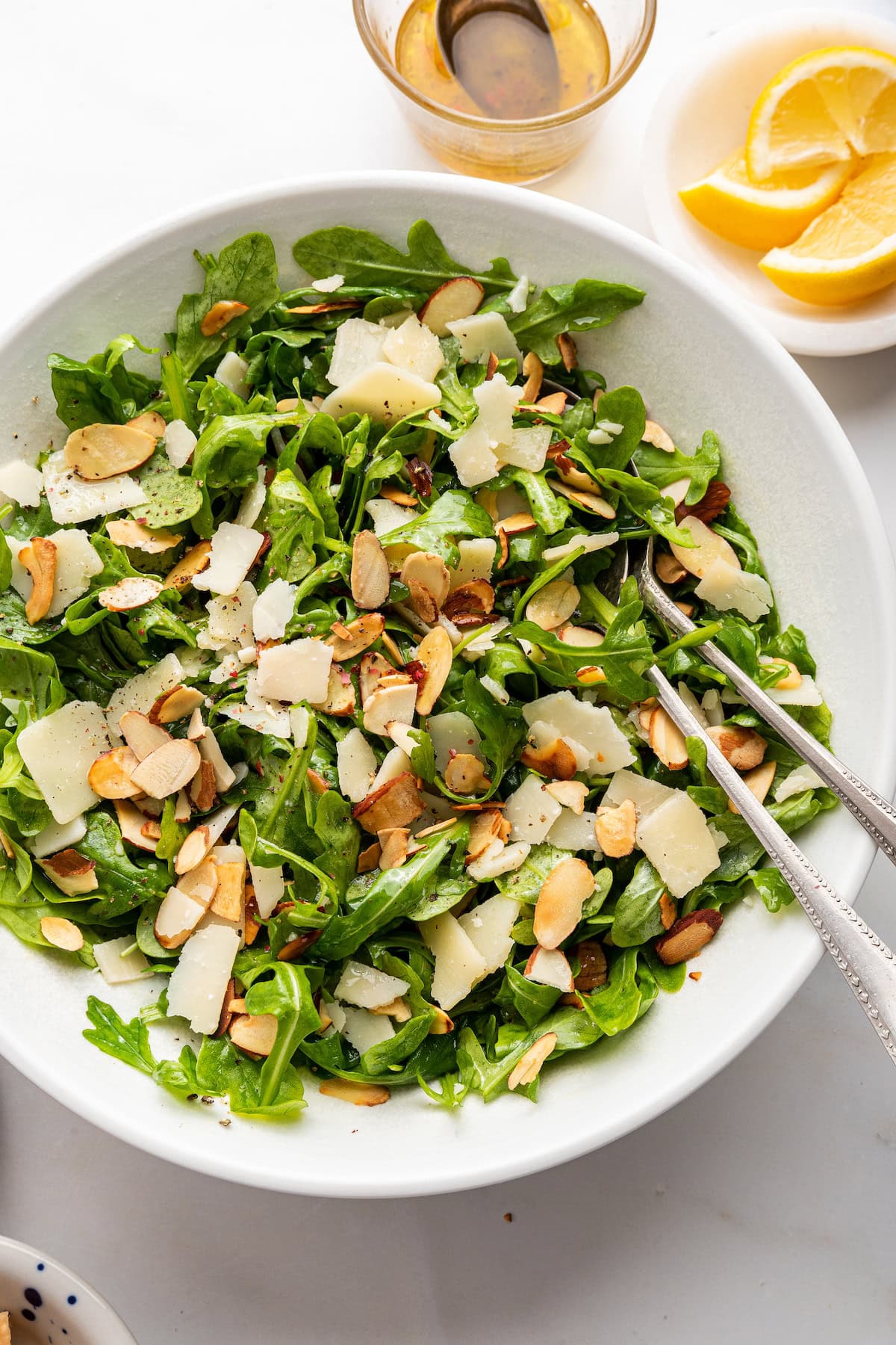 Arugula salad served in a white bowl with silver serving spoons.