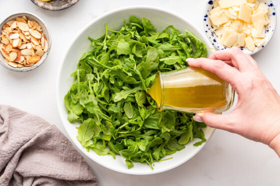 A woman's hand pouring the dressing over the arugula.