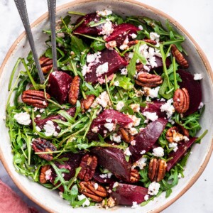 A beet salad with arugula, pecans, and crumbled feta in a bowl with two metal serving spoons.