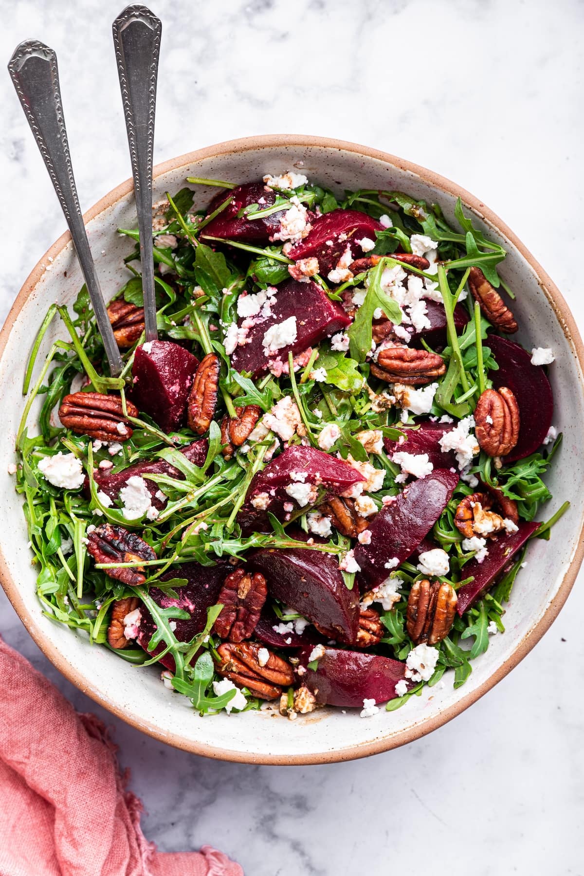 A beet salad with arugula, pecans, and crumbled feta in a bowl with two metal serving spoons.