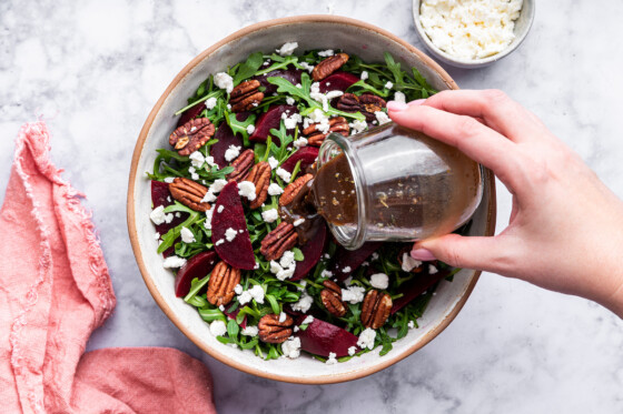 Dressing being poured into a beet salad with arugula, pecans, and crumbled feta in a bowl.