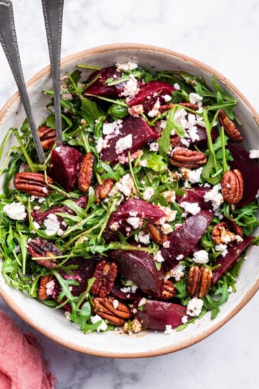A beet salad with arugula, pecans, and crumbled feta in a bowl with two metal serving spoons.