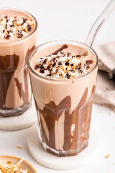 A samoa cookie protein shake in a glass topped with coconut and drizzled chocolate. The glass has drizzled chocolate around the sides.