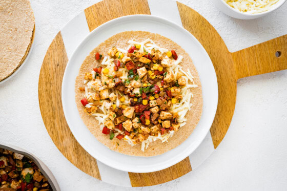 A large whole wheat tortilla on a plate topped with cheese, chicken, corn, tomato, and onion.
