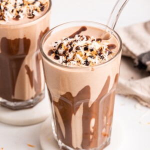 A samoa cookie protein shake in a glass topped with coconut and drizzled chocolate. The glass has drizzled chocolate around the sides.