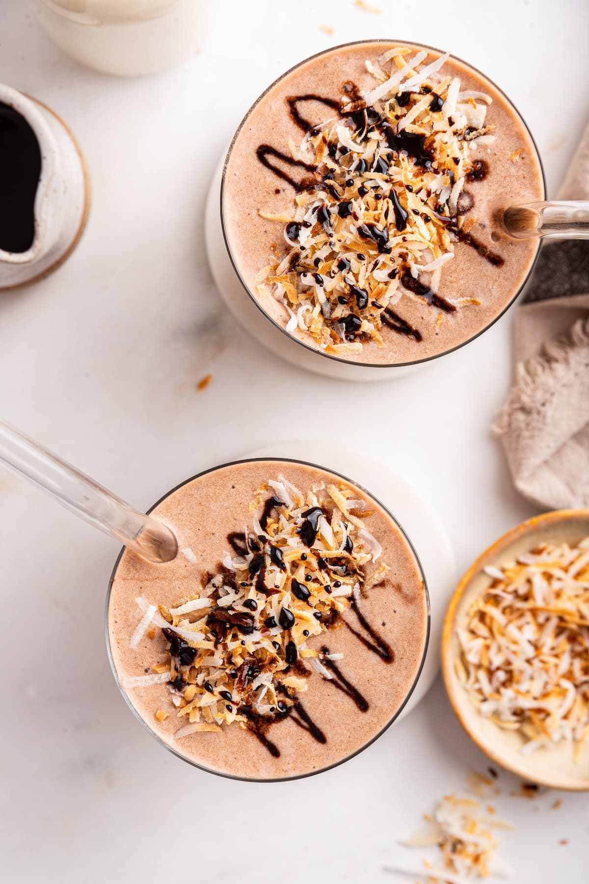 Two samoa cookie protein shakes in a glass cups with coconut shreds and a chocolate drizzle topping.