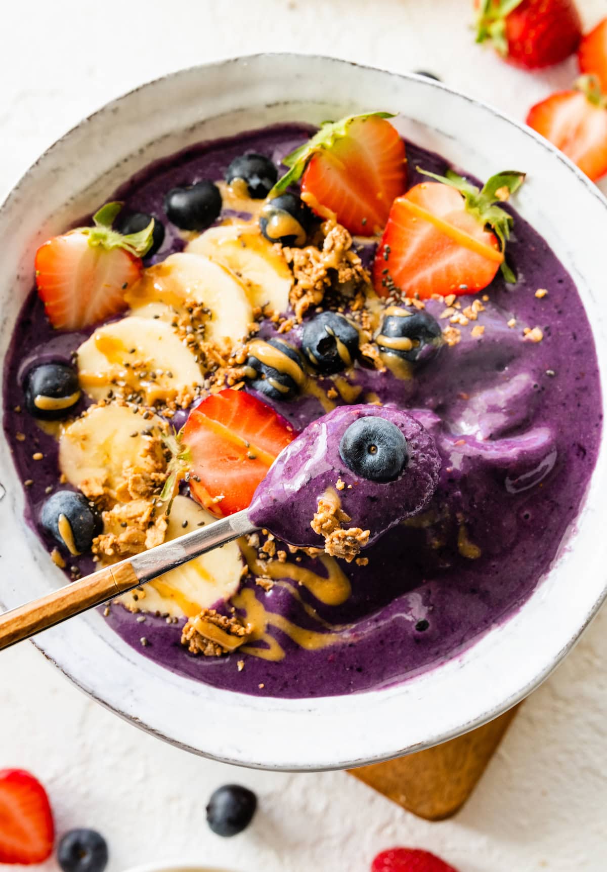 A spoon holds a bite size portion of a smoothie bowl. The bowl is topped with fresh fruit, granola, and a drizzle of almond butter.