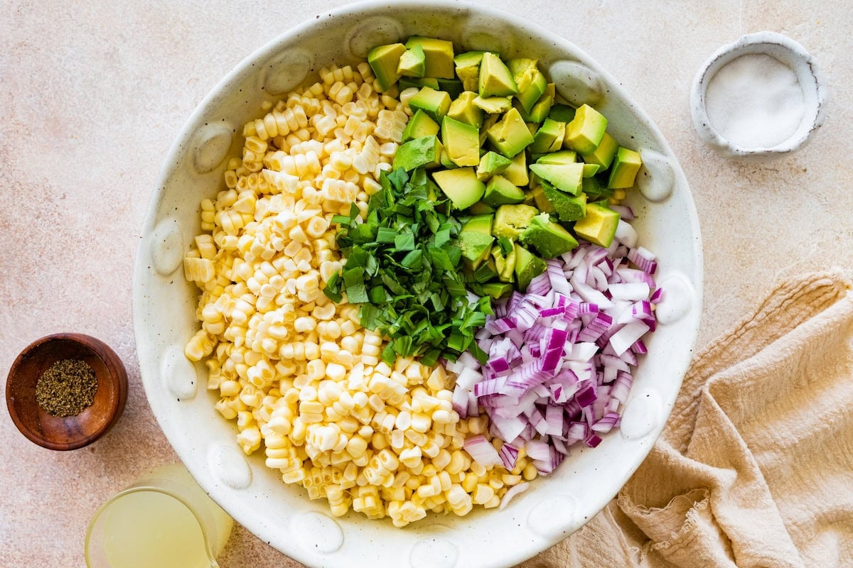 Ingredients used for an avocado corn salad in a large bowl. These ingredients include corn, cubed avocado, red onion, and basil.